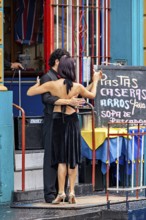 A couple dances tango in front of a colourful building, hugging each other, next to a menu board,