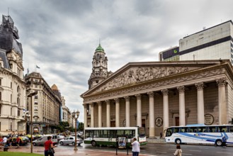An impressive columned building against an overcast sky, surrounded by city buses, Historic