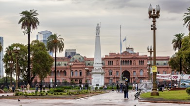 A monument on a square with a historic pink building and palm trees in the background under a
