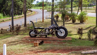 A metal motorbike bust stands in a park with green spaces and trees, placed on a pedestal, A