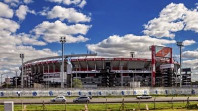 A modern stadium with stands and floodlights under a clear sky and passing cars, the River Plate