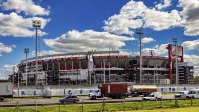 A large stadium with several stands and floodlights under a blue sky and passing vehicles, the