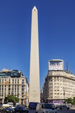 A tall obelisk stands between modern buildings under a clear blue sky, The Great Obelisk in the