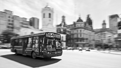 A bus drives through a busy city with historic buildings in the background in black and white, Bus