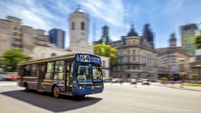 A colourful bus drives through a busy city with historic buildings and a blue sky in the