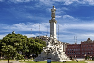 A large monument with a statue in front of trees and historic buildings under a blue sky, The city