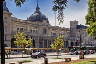 A historic railway station with a domed roof, surrounded by cars and trees in daylight, The city of