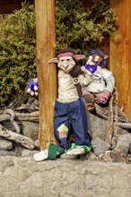 Two dolls lean against tree trunks next to a stone wall, decorated in a rustic setting, dwarves and