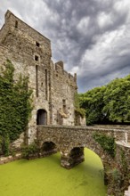 Old stone castle with overgrown wall and bridge over a green moat under a cloudy sky, Pirou Castle