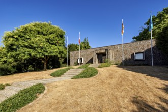 A modern building with flags, surrounded by trees and a dry meadow under a clear sky, German