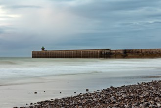 A breakwater juts into the sea, surrounded by a pebble beach and a cloudy sky, The pier and beach
