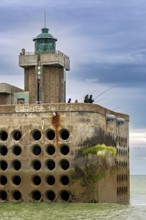 Concrete building with lighthouse and anglers at the water's edge, under a cloudy sky, The harbour