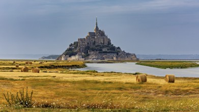 A castle rises in the middle of fields and a river, surrounded by bales of straw, Mont Saint Michel