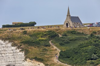 A small church stands on grassy cliffs with a path and walkers, under a blue sky, The church of