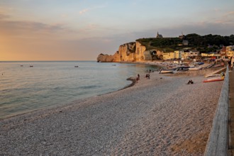 Beach with pebbles and boats, surrounded by rocks and a village, in the warm light of the sunset,