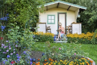 A woman sits relaxed on the small veranda of a garden shed and reads. Blooming summer flowers in