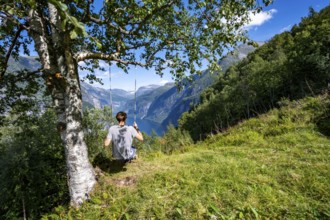 Young man swinging on a tree, view of idyllic fjord landscape of the Geirangerfjord at Blomberg