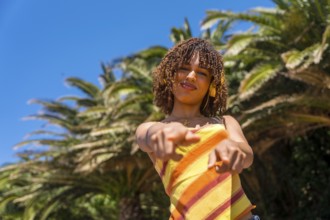 Stylish young woman wearing headphones points directly at the camera in front of palm trees in a