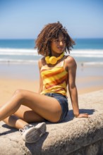 Young woman with curly hair and yellow headphones enjoys the beach view while sitting on a concrete