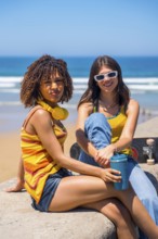Two young women relaxing on beach wall, enjoying ocean view with skateboard and reusable coffee