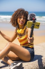Young woman vlogger recording video blog on sunny beach using smartphone, gimbal and microphone