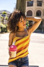 Stylish young woman with curly hair enjoys a cold beverage on a sunny summer day, exuding a