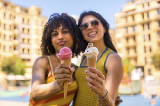 Two young women smiling brightly while holding colorful ice cream cones on a sunny summer day in