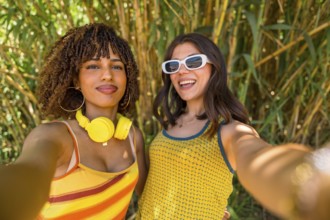 Two cheerful young women with colorful clothes and headphones taking a selfie in front of a bamboo