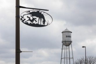 Bandera water tower behind artistic metal sign showing cowboy life on pole in Bandera, Texas, USA