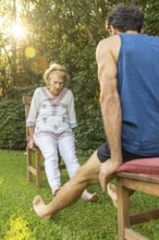 Senior woman exercising her legs with the help of a personal trainer, sitting on chairs in a
