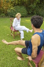Elderly woman exercising her legs with the help of a physiotherapist, sitting on chairs in a