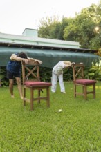 Senior woman and her physiotherapist are bending forward, holding onto chairs in a garden,