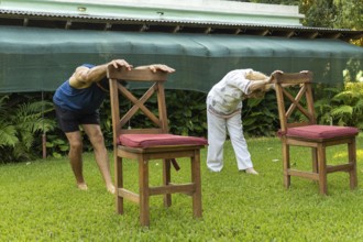 Elderly man and woman practicing stretching exercises with chairs on a grassy lawn in a serene