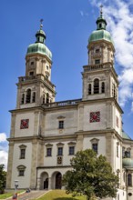 Baroque church with two towers and clocks against a blue sky, surrounded by trees, The town of