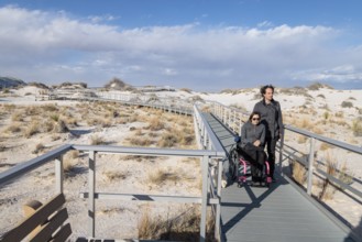 Man and handicapped woman in wheelchair stroll down the Interdune Boardwalk with their ped dog at