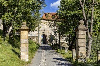 Entrance gate to the castle, flanked by stone pillars and trees, under a bright blue sky, The