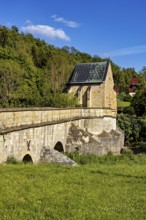 An old stone bridge with a small chapel in the background, surrounded by green trees under a blue