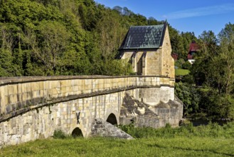 Side view of a historic stone bridge with a chapel and lush vegetation in the background, The
