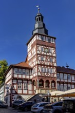 Half-timbered tower with clock surrounded by historic buildings and parked cars on a clear day, The