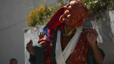 A masked dancer in shades of orange and red with flowers in the background in Leh, trekking in