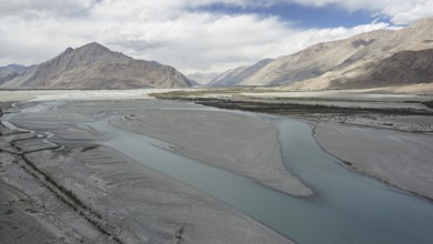 A calm river, Indus flows through a vast landscape surrounded by mountains under a cloudy sky,