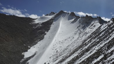 Snowy mountain peak at Khardung-la Pass under clear blue sky with some clouds and rocky landscape,