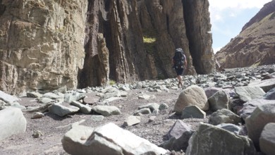 A hiker with a backpack walks through a rocky gorge under a blue sky, trekking at Stok La Pass in