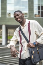 Young black man smiling and posing with laptop bag on his shoulder, leaning against a railing