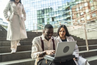 Three diverse business people collaborating outdoors, two using a laptop on city stairs while a