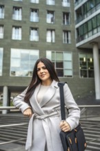Confident businesswoman wearing a coat and carrying a shoulder bag, standing outdoors in an urban