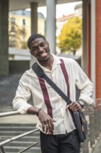 Smiling young black student on a university campus, leaning on a railing with a shoulder bag,
