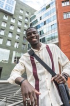 Young black man smiling and leaning on a railing in a modern city street, carrying a bag and