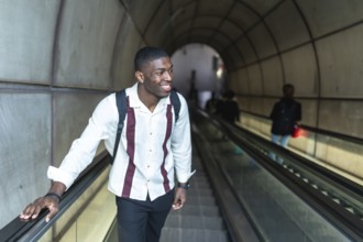 Young black man smiling while traveling up an escalator in a subway station tunnel, carrying a