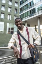 Confident young black professional smiling at camera, leaning on a railing with a messenger bag in